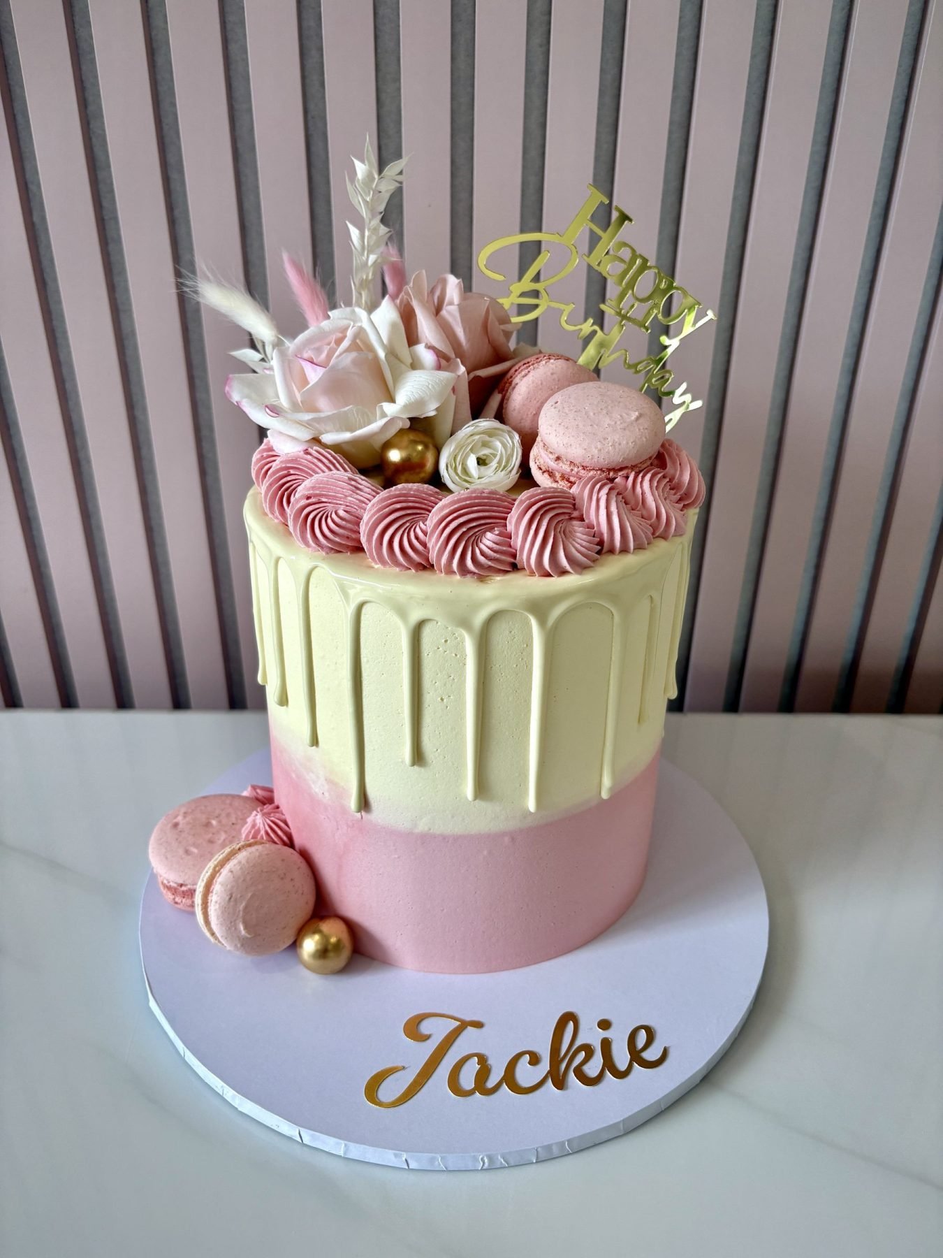 Pastel pink and ivory drip cake decorated with macarons, blush roses, buttercream swirls and gold accents.